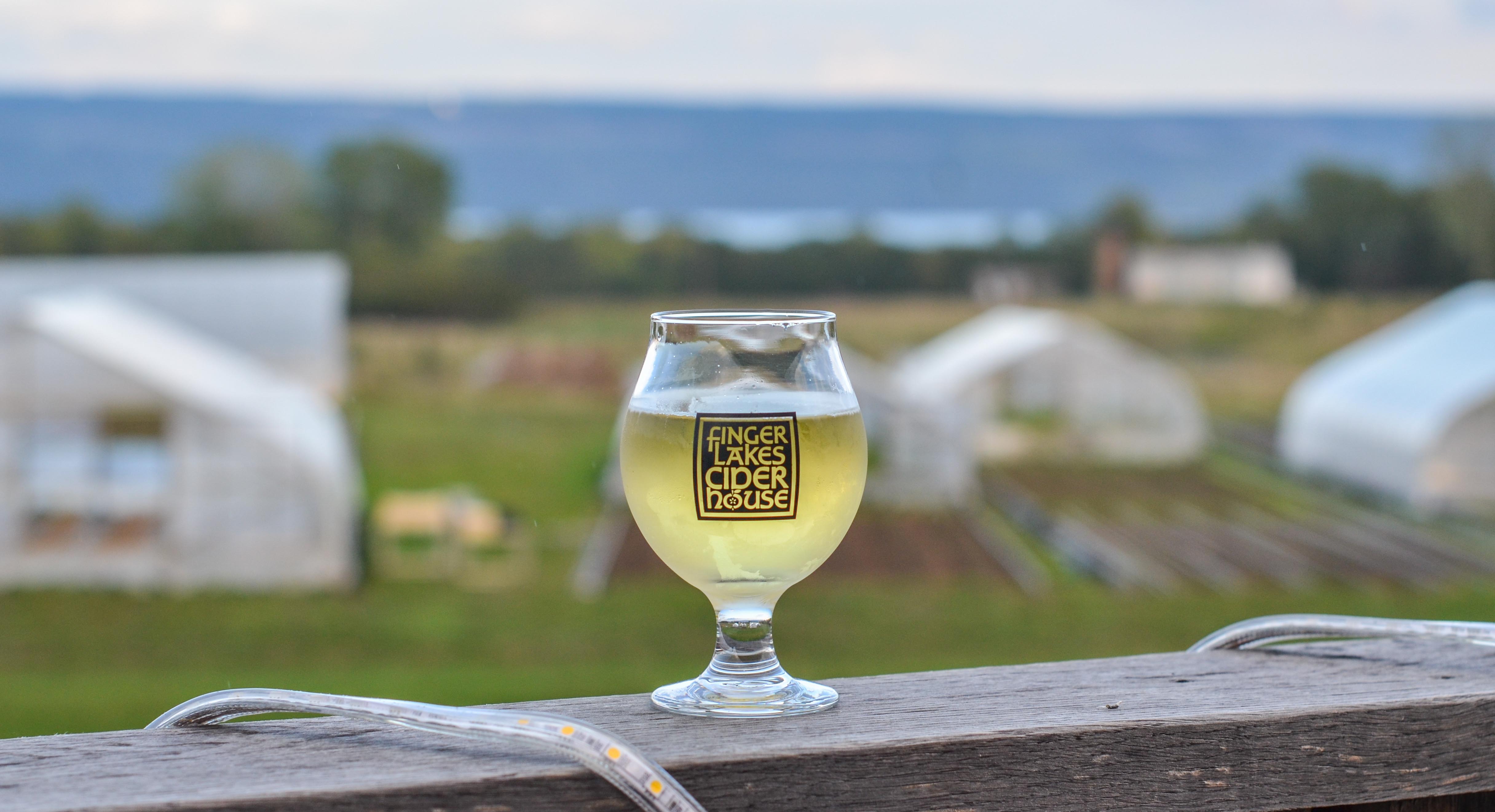 Finger Lakes Cider House cider glass with Farmhouse Brewery fields and greenhouses in background, Finger Lakes region NY
